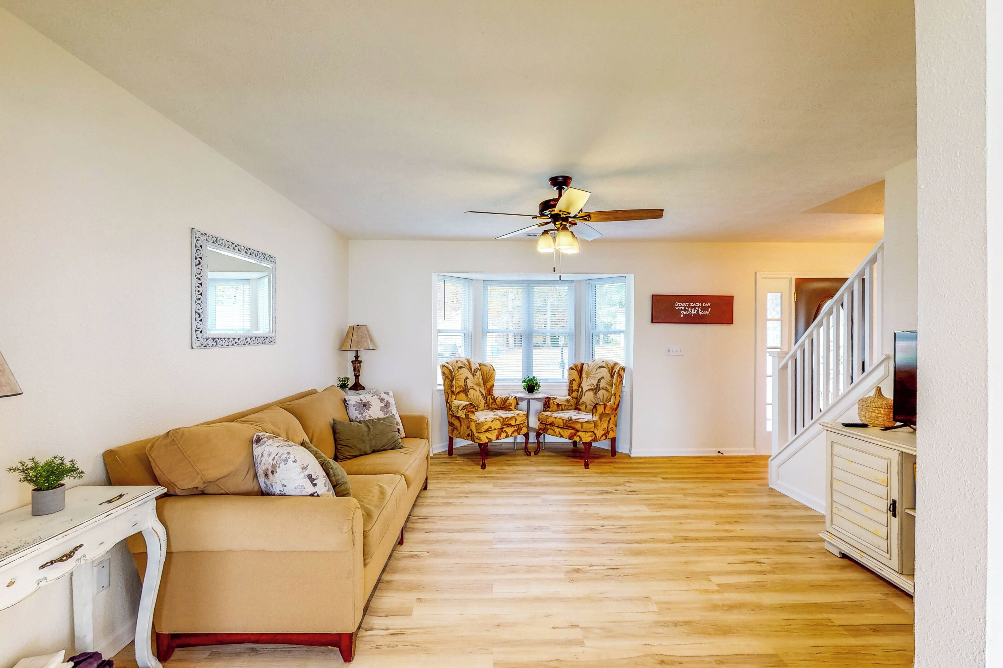 A living room with a beige sofa, two armchairs, a ceiling fan, and a small table with a lamp.