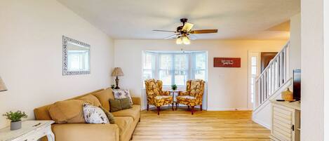 A living room with a beige sofa, two armchairs, a ceiling fan, and a small table with a lamp.