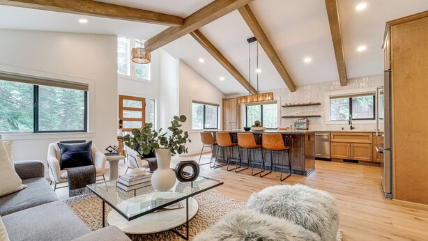 A spacious living room with a sofa, coffee table, and a large window. A kitchen with wooden cabinets and a bar area is visible in the background. - Walk to Gondola: Truckee Retreat w/ Resort Perks! (Truckee)