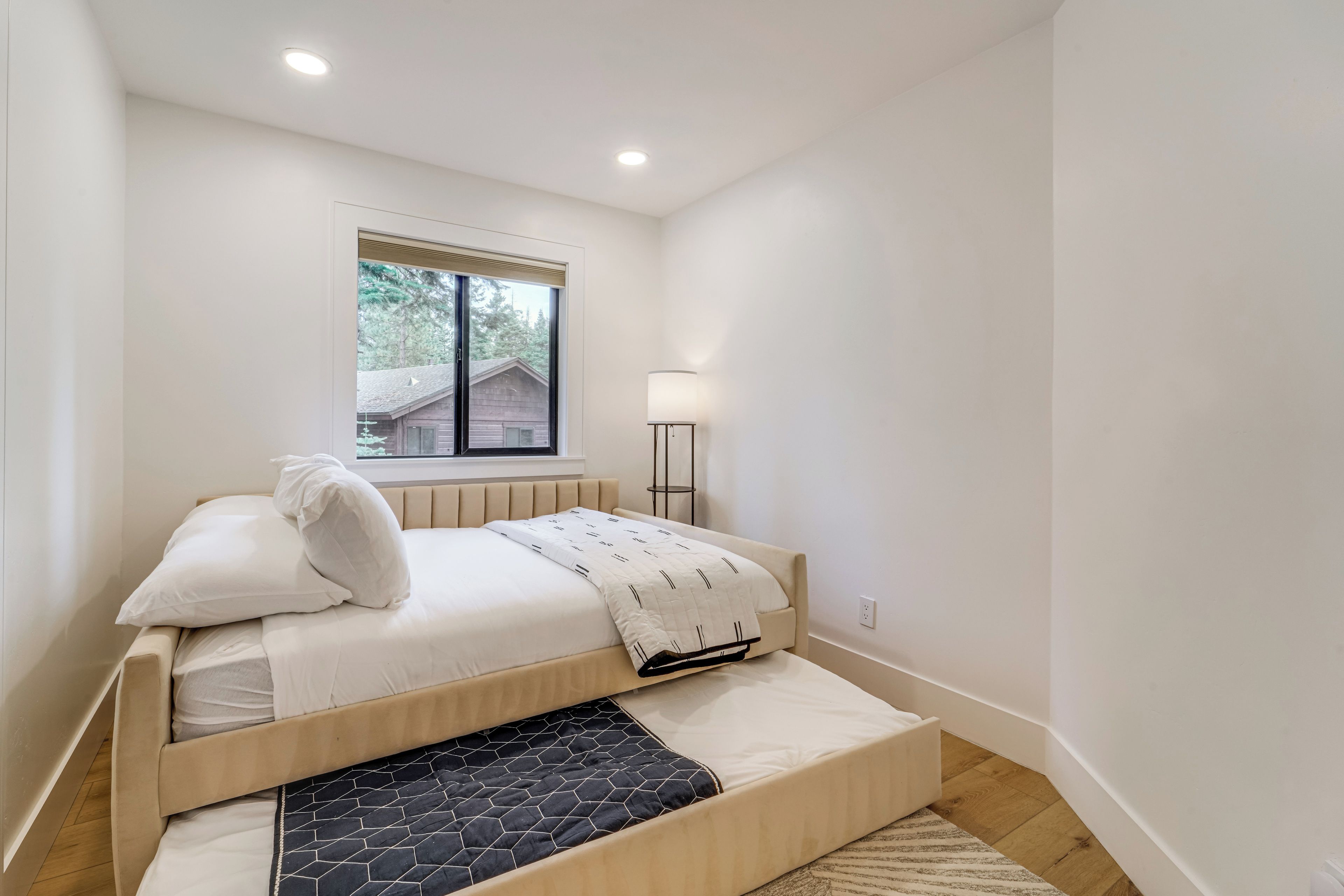 A bedroom with a bed, a window, a lamp, and a view of a house outside.