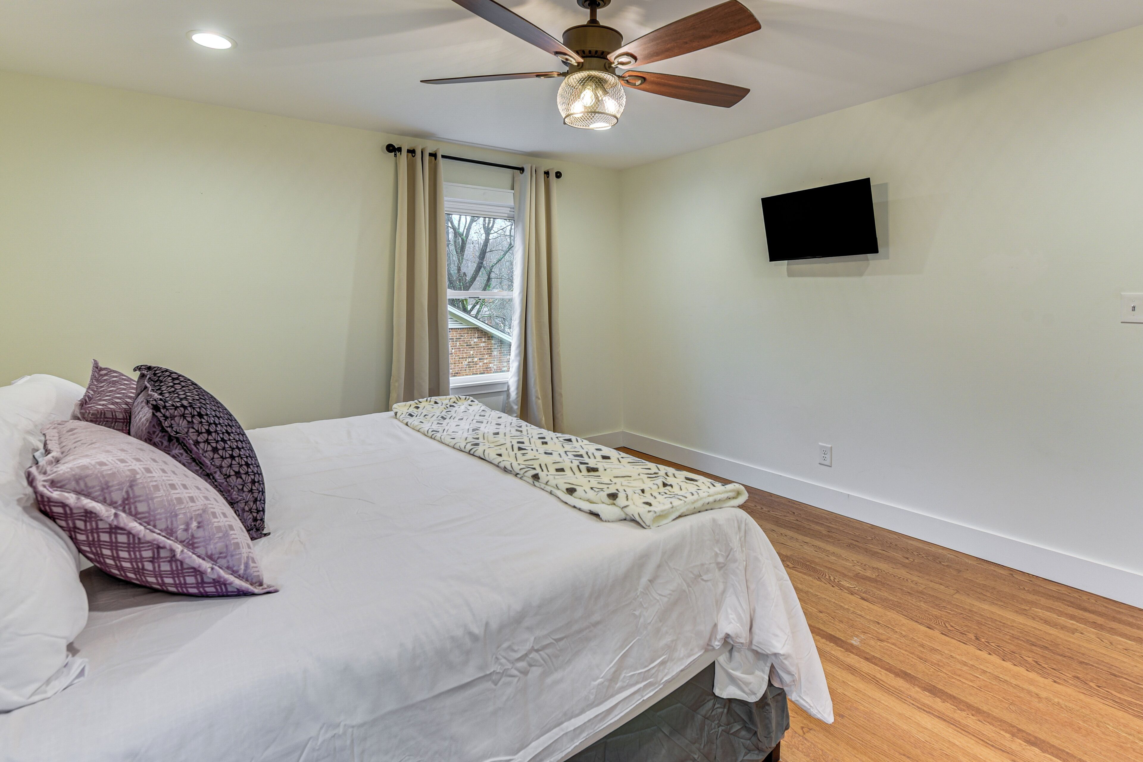A bedroom with a bed, pillows, a ceiling fan, a window with curtains, and a mounted TV.