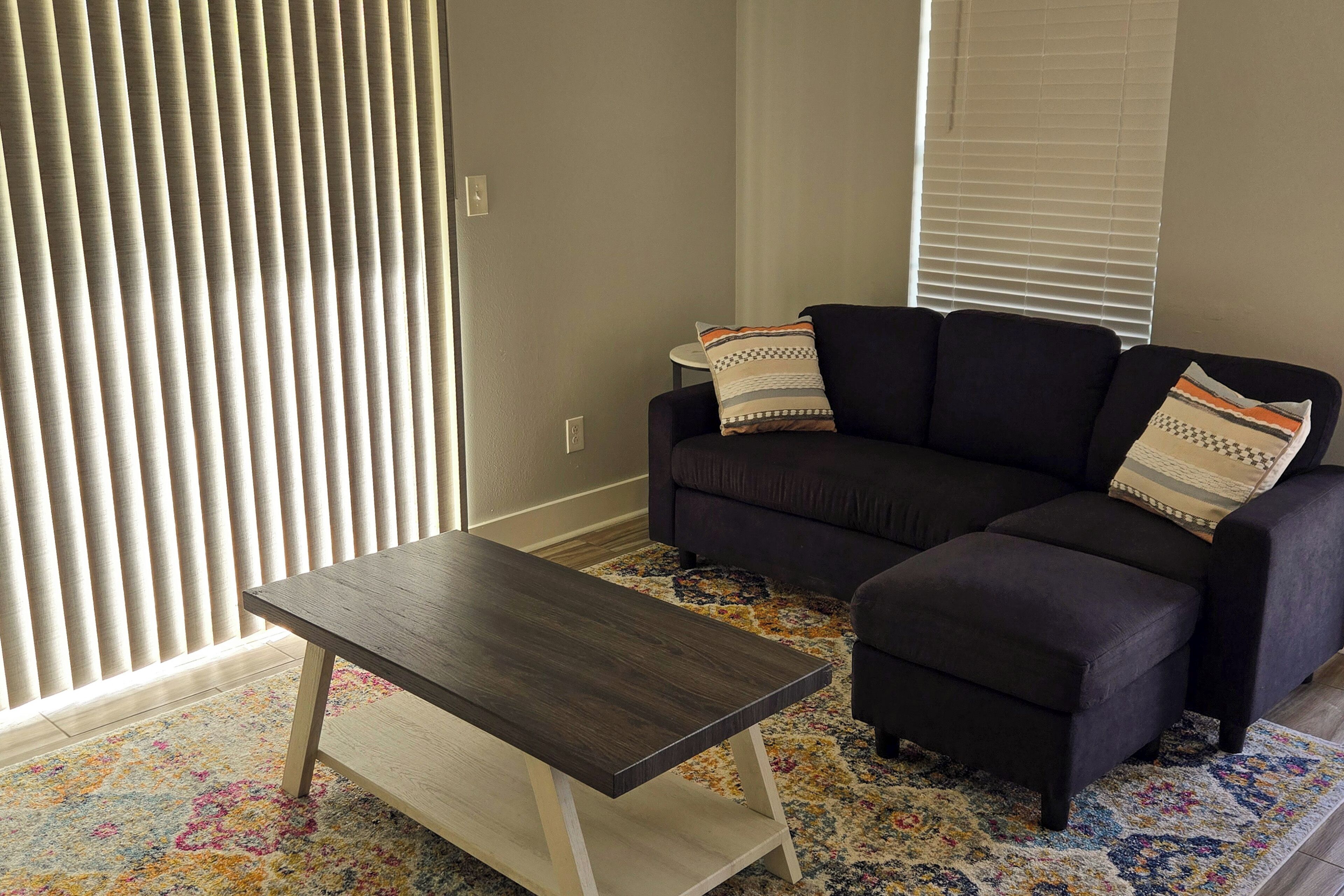 A living room with a dark sofa, a matching ottoman, a wooden coffee table, and a colorful rug.
