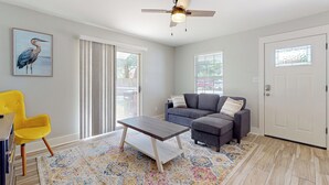 A living room with a gray sofa, a yellow chair, a coffee table, a ceiling fan, and a large window with blinds. - Cozy Duplex Spacious Close to Everything (Pensacola)