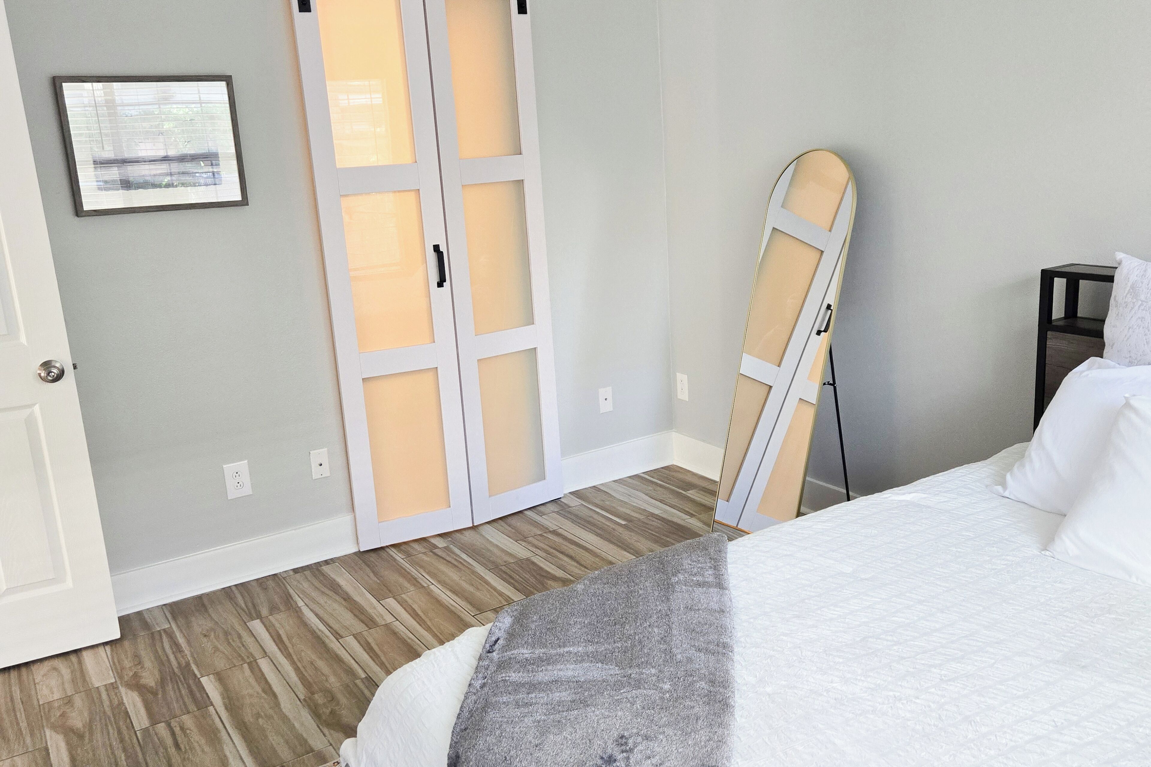 A bedroom with a bed, a wooden floor, a wardrobe with frosted glass doors, a mirror, and a nightstand.