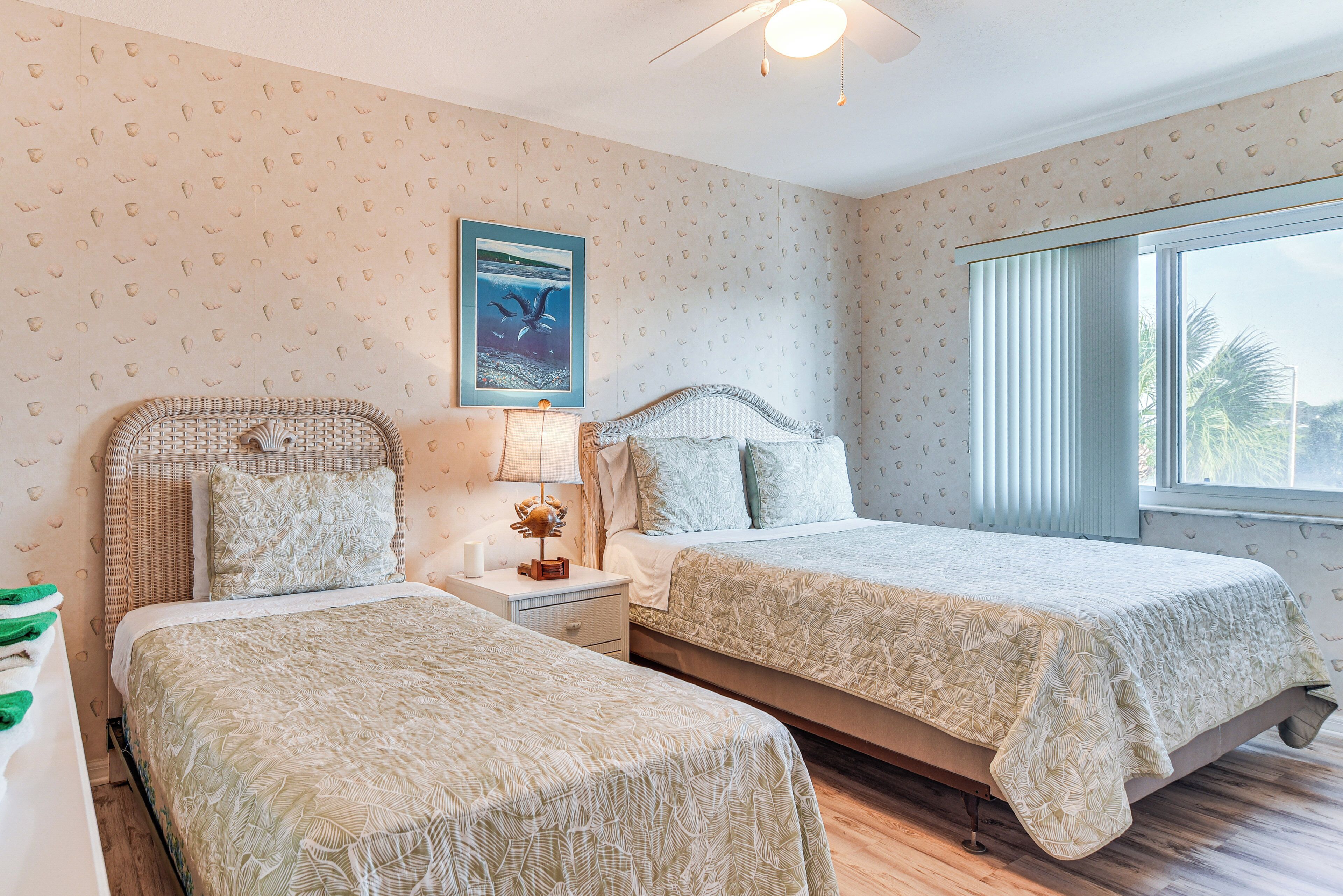 A bedroom with two beds, a nightstand with a lamp, a framed picture on the wall, and a window with blinds.