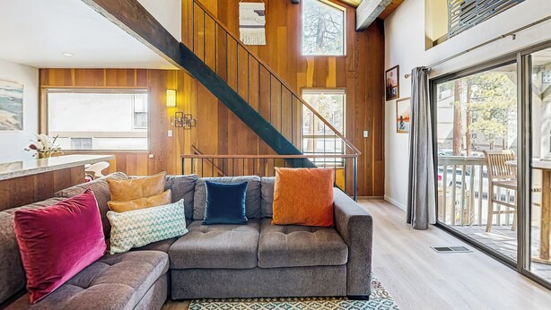 A modern living room with a gray sectional sofa, a wooden staircase, and large windows offering views of the outdoors. - Lake Tahoe Adventure Base! Townhome w/ Pool Access (Kings Beach)