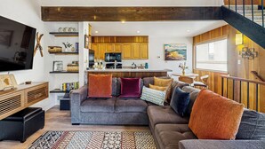 A modern living room with a grey sofa, wooden beams, a kitchen in the background, and a staircase. - Lake Tahoe Adventure Base! Townhome w/ Pool Access (Kings Beach)