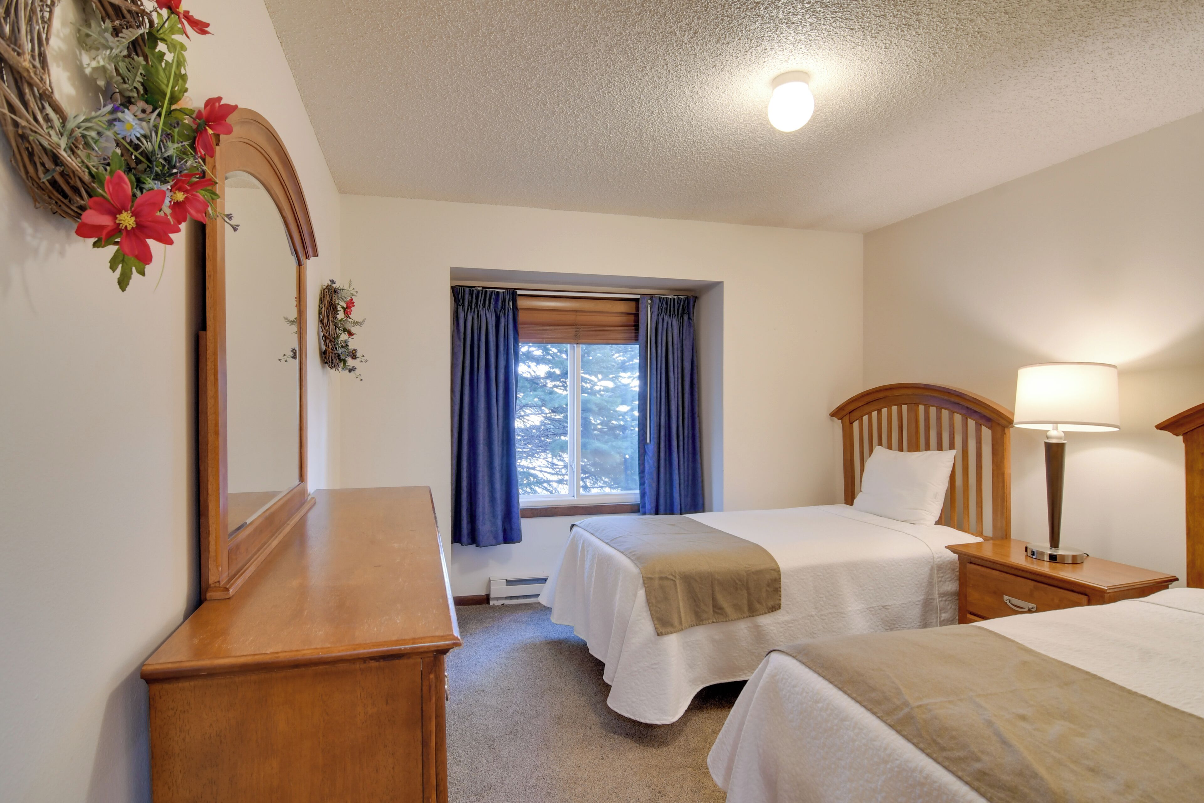 A hotel room with two beds, a wooden dresser, a mirror, a window with blue curtains, and a wall-mounted light fixture.