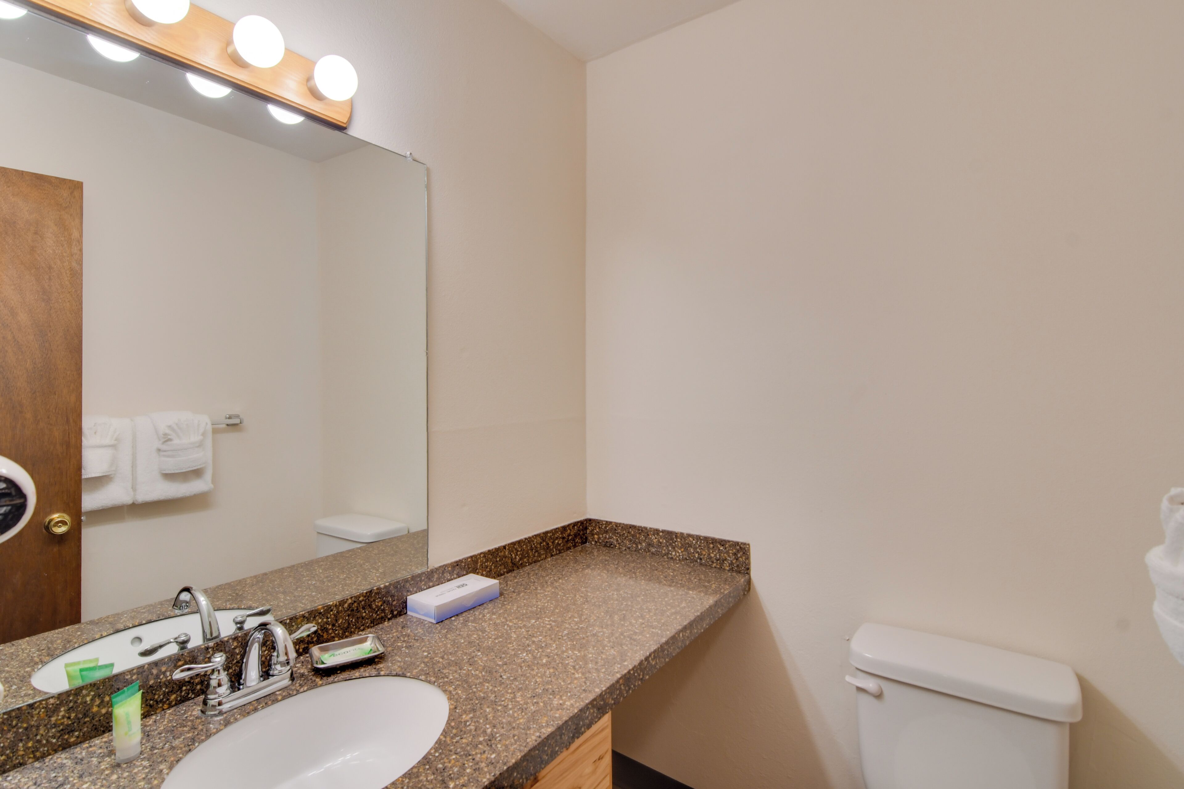 A bathroom with a granite countertop, a sink, a mirror with lights, a toilet, and a towel rack.