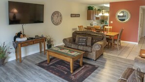 Sala de estar con sofá, mesa de centro y televisor. Hay una zona de comedor con mesa y sillas. El piso es de madera y se ve una cocina al fondo.