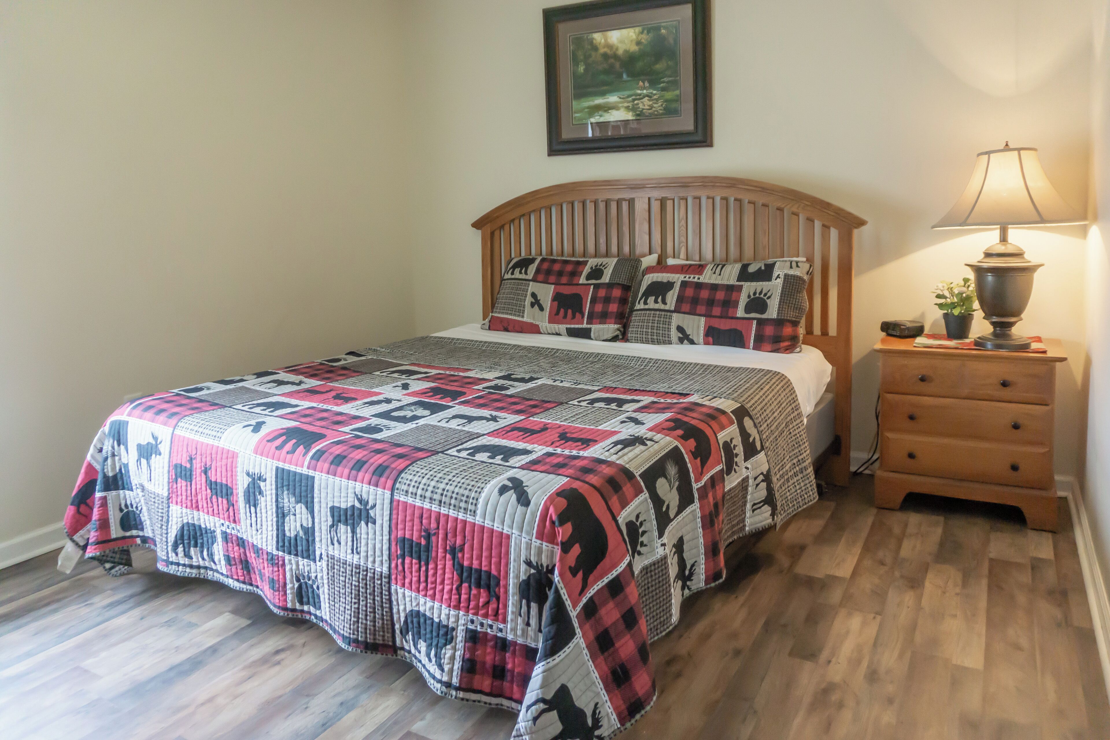 A bed with a quilt featuring animal patterns, a wooden headboard, a bedside table with a lamp, and a framed picture on the wall.