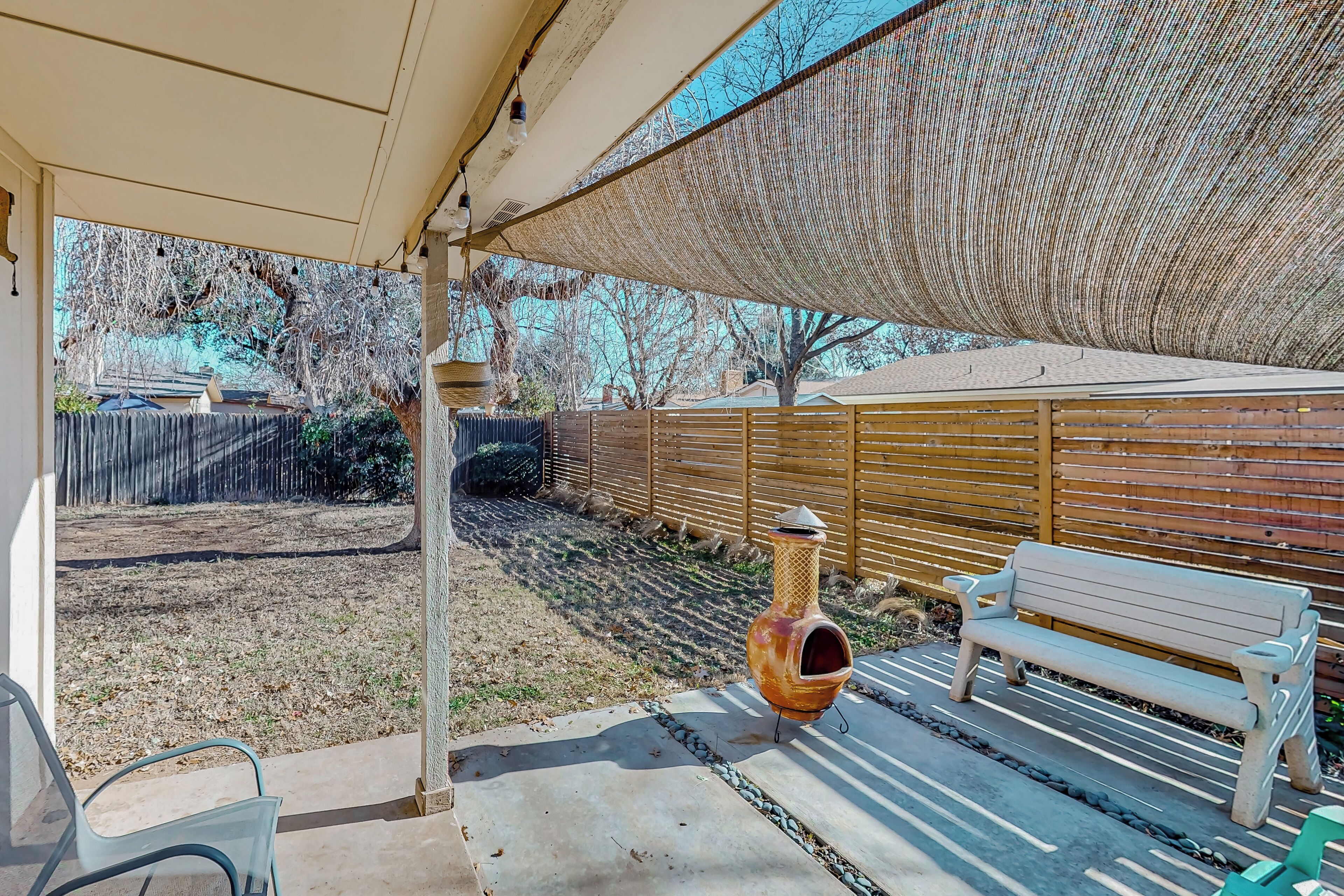 A covered patio with a wooden bench, a decorative urn, and a chain-link fence in the background.