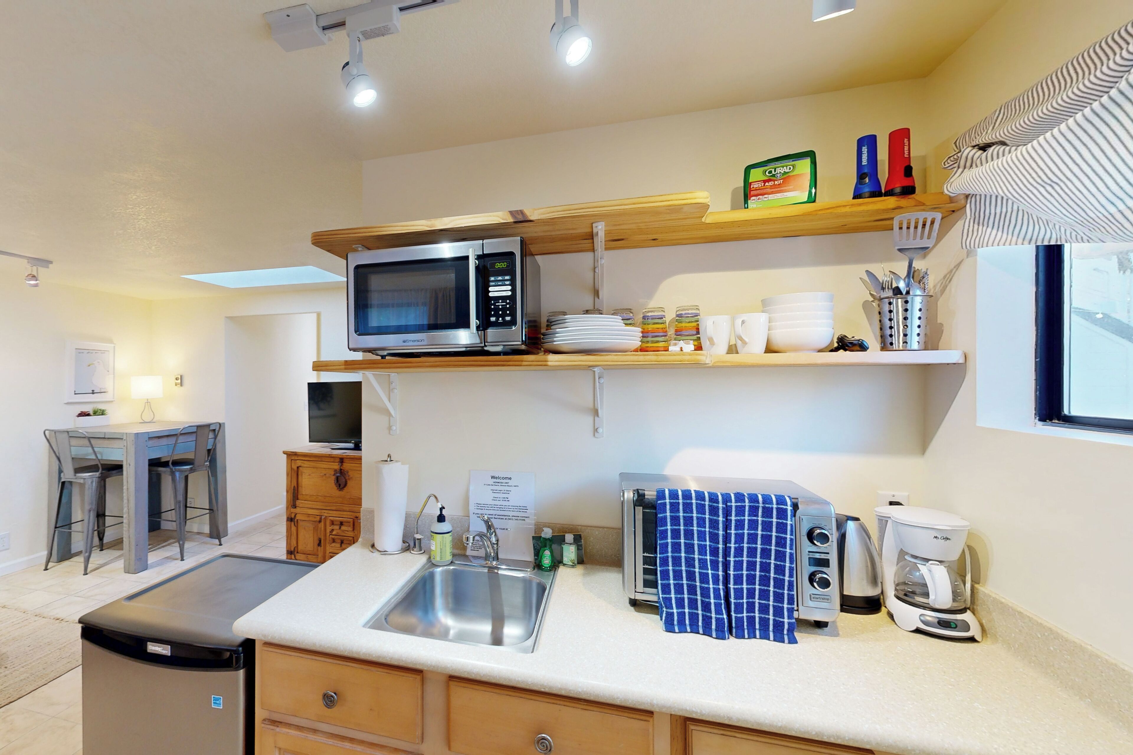 A compact kitchen with a microwave, toaster, coffee maker, and a sink. There are wooden cabinets and a small dining area with a table and chairs.