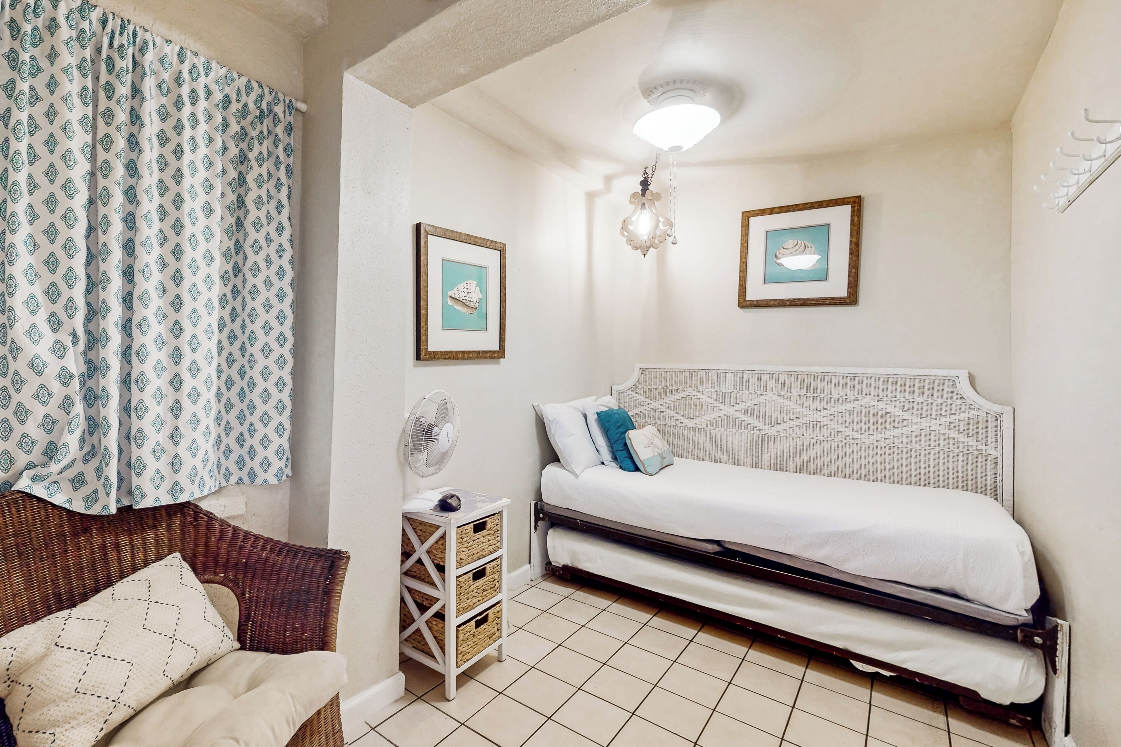 Ein kleines, gemütliches Schlafzimmer mit einem Einzelbett, einem Korbsessel, einem Nachttisch mit Ventilator und zwei gerahmten Bildern an der Wand.