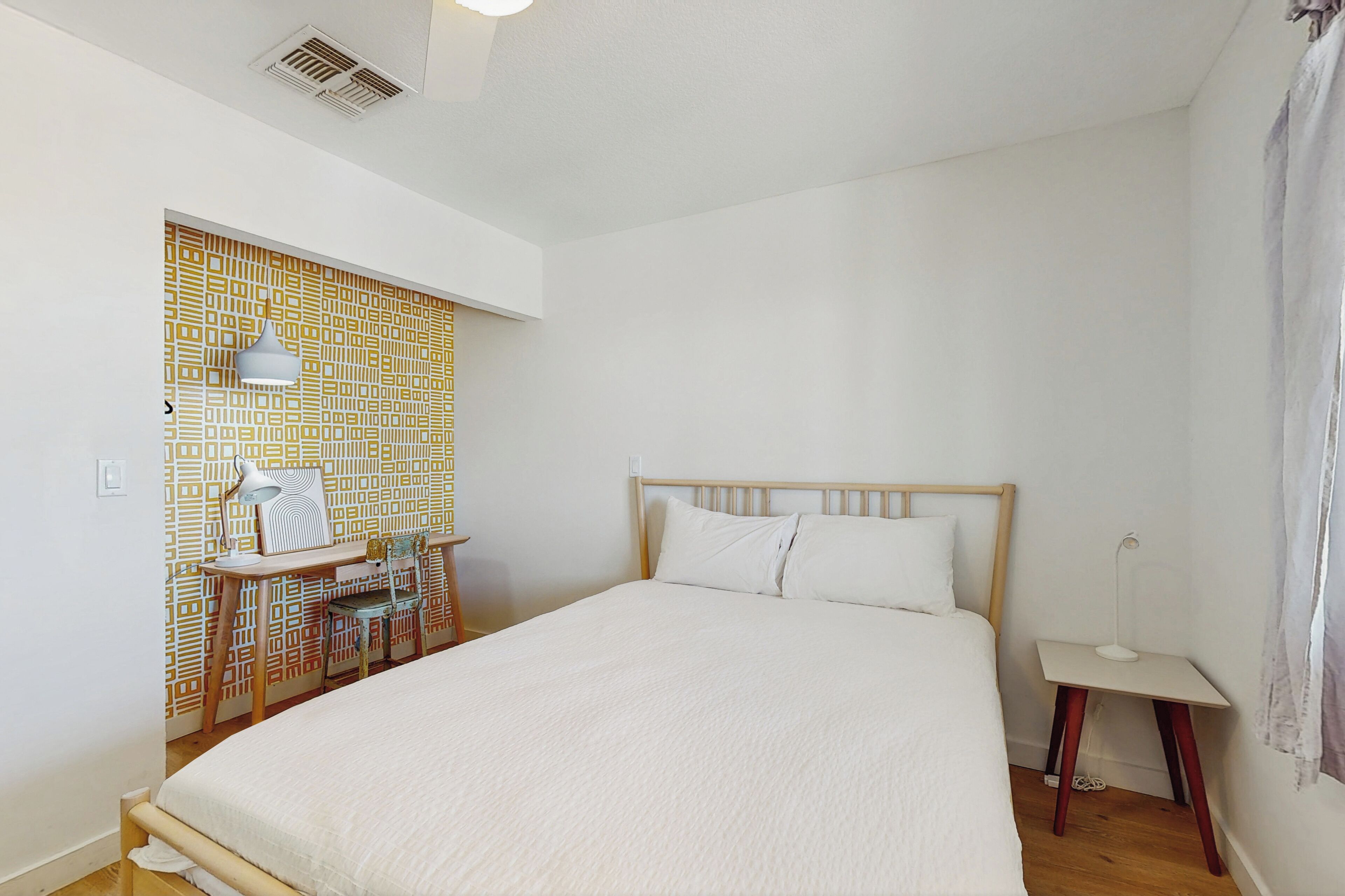 A bedroom with a large bed, a desk with a chair, a lamp, and a patterned wall.
