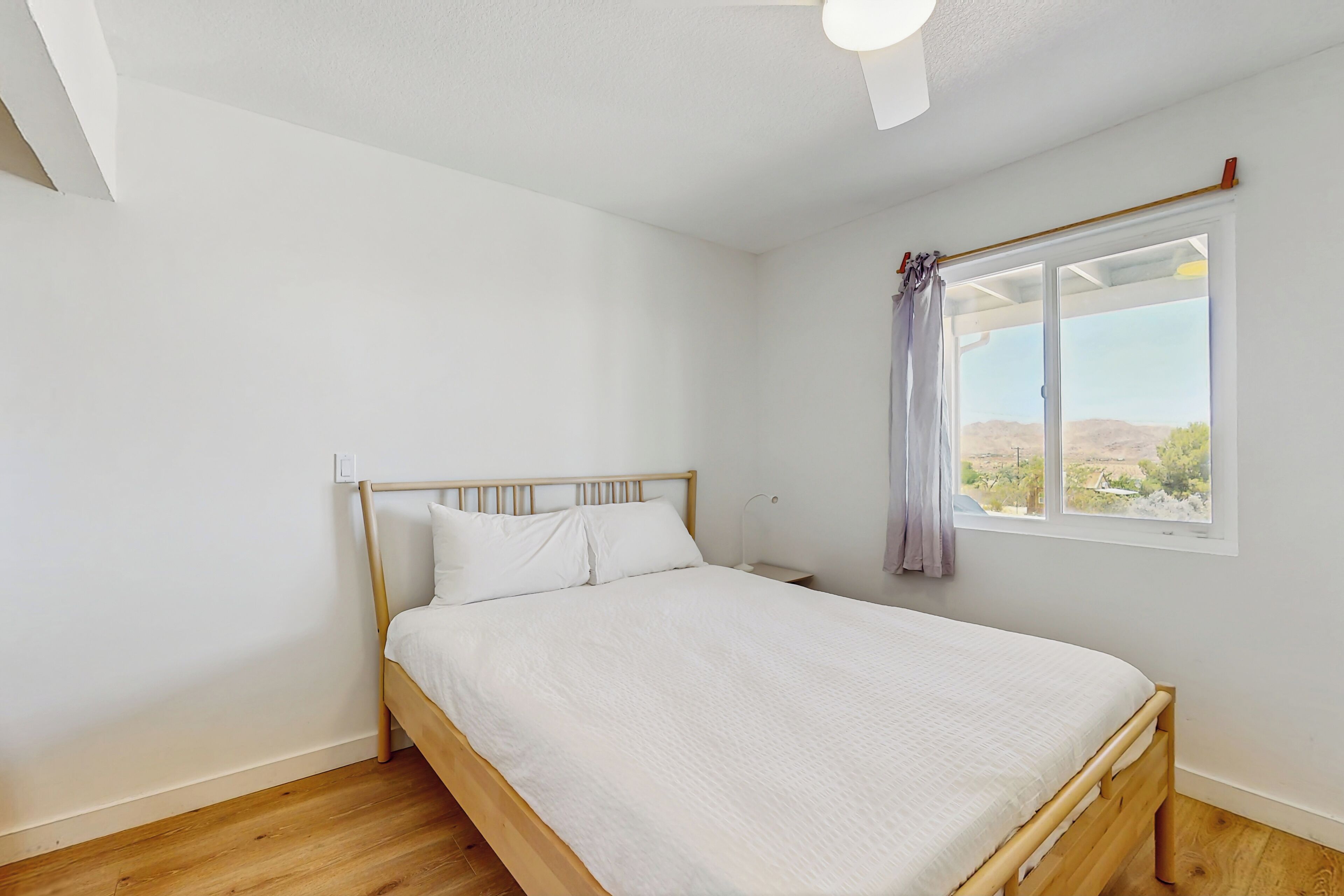 Une chambre avec un lit en bois, des murs blancs, une fenêtre donnant sur l’extérieur et un ventilateur de plafond.