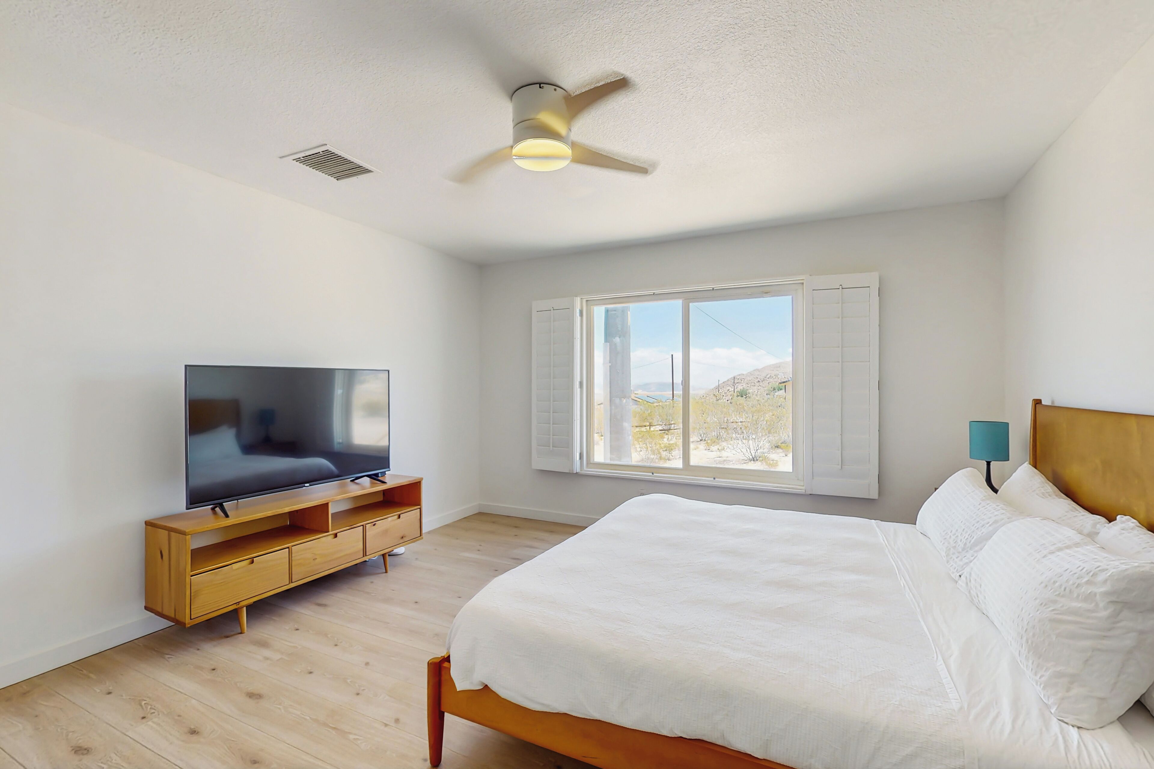 Une chambre à coucher avec un lit, un téléviseur posé sur un meuble en bois, un ventilateur de plafond, une fenêtre avec des volets et une table de chevet avec une lampe.