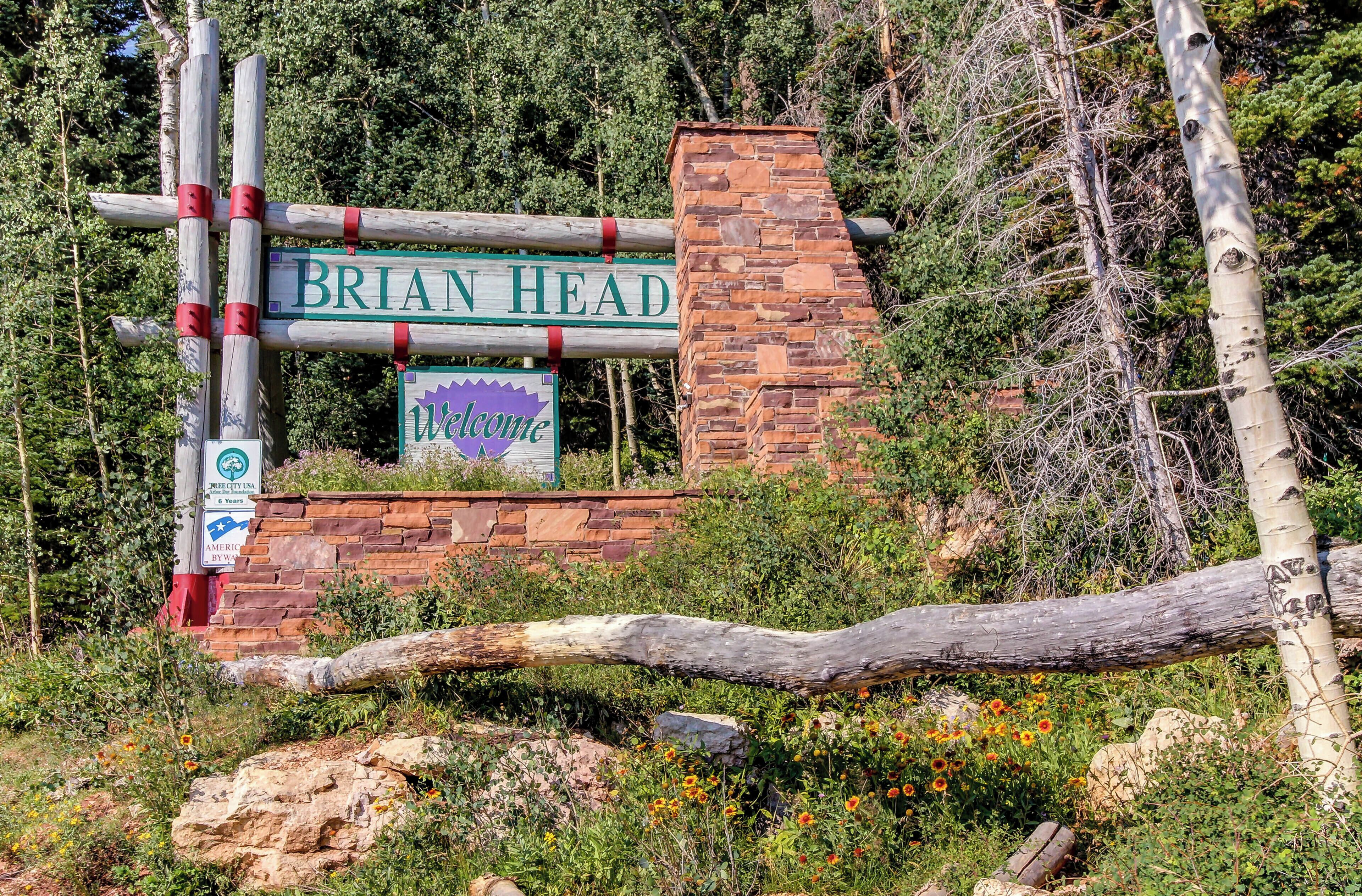 A sign reading "Brian Head" with a welcome message, set against a backdrop of trees and a stone wall.