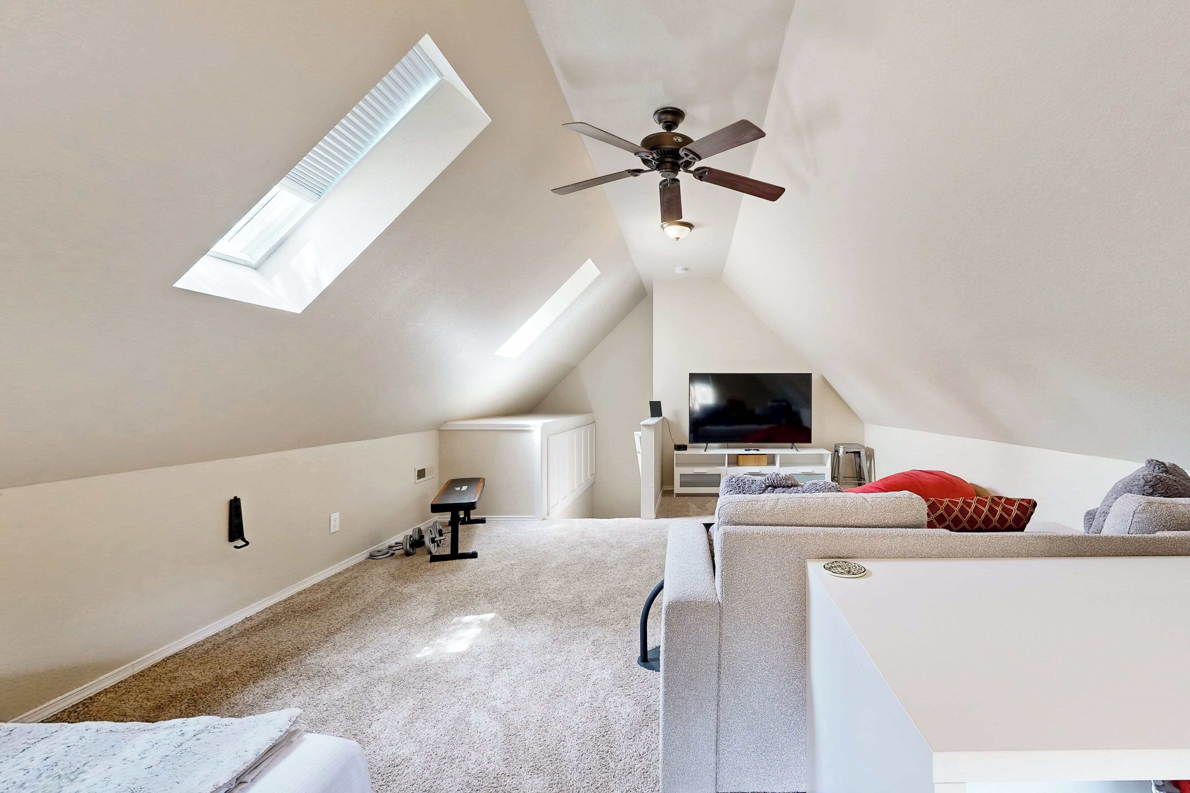 A high-ceilinged attic bedroom with a ceiling fan, a large window, a flat-screen TV, and a cozy seating area.