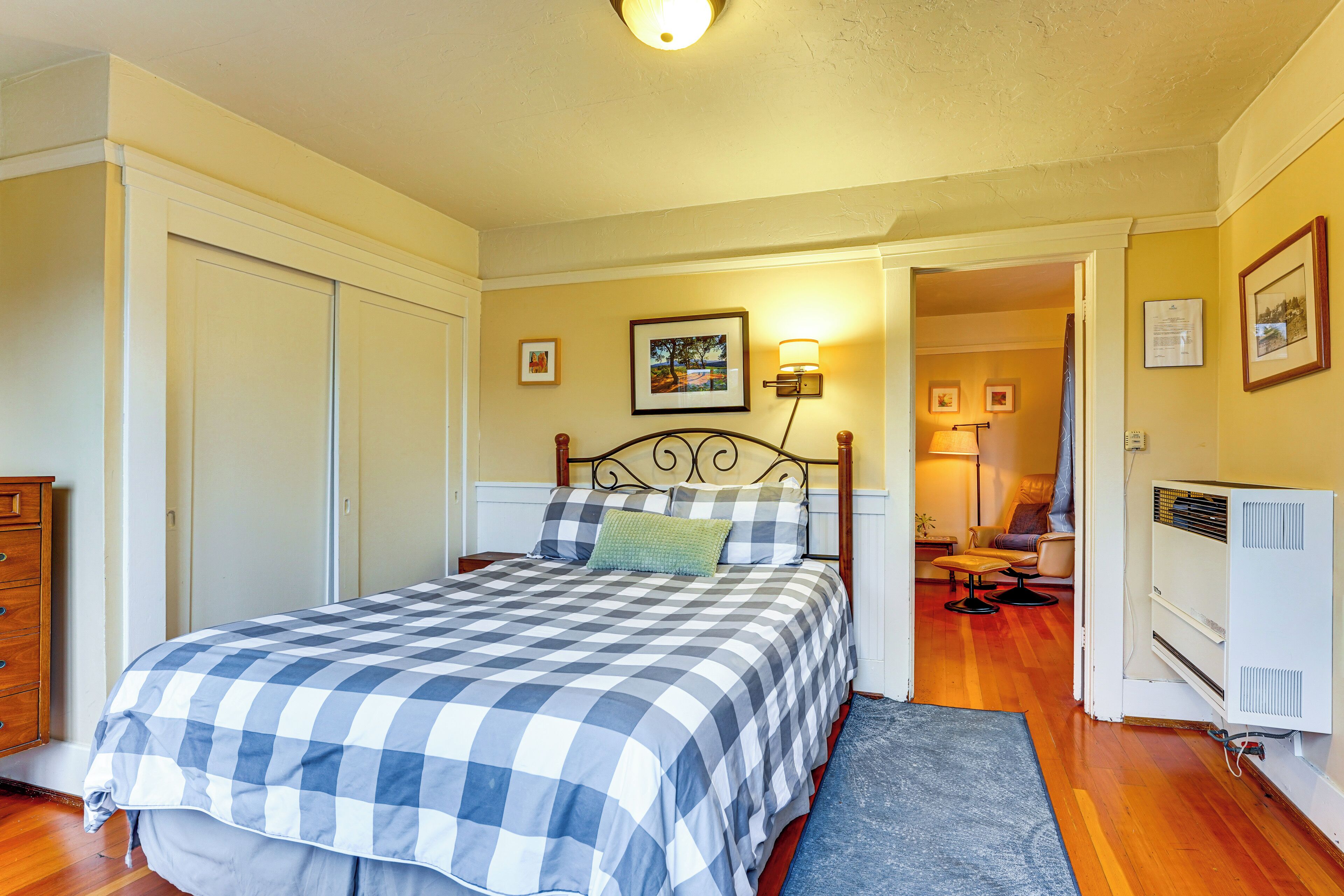 A bedroom with a checkered bedspread, a wooden dresser, a radiator, and a door leading to another room.