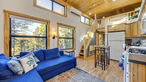Interior de una cabaña moderna con un sofá azul, ventanas amplias, una cocina y una escalera.
