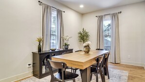 A dining area with a wooden table, six chairs, a dark console table with a vase and plants, and two windows with curtains. - Game Room & Backyard: Modern Altoona Home! (Altoona)