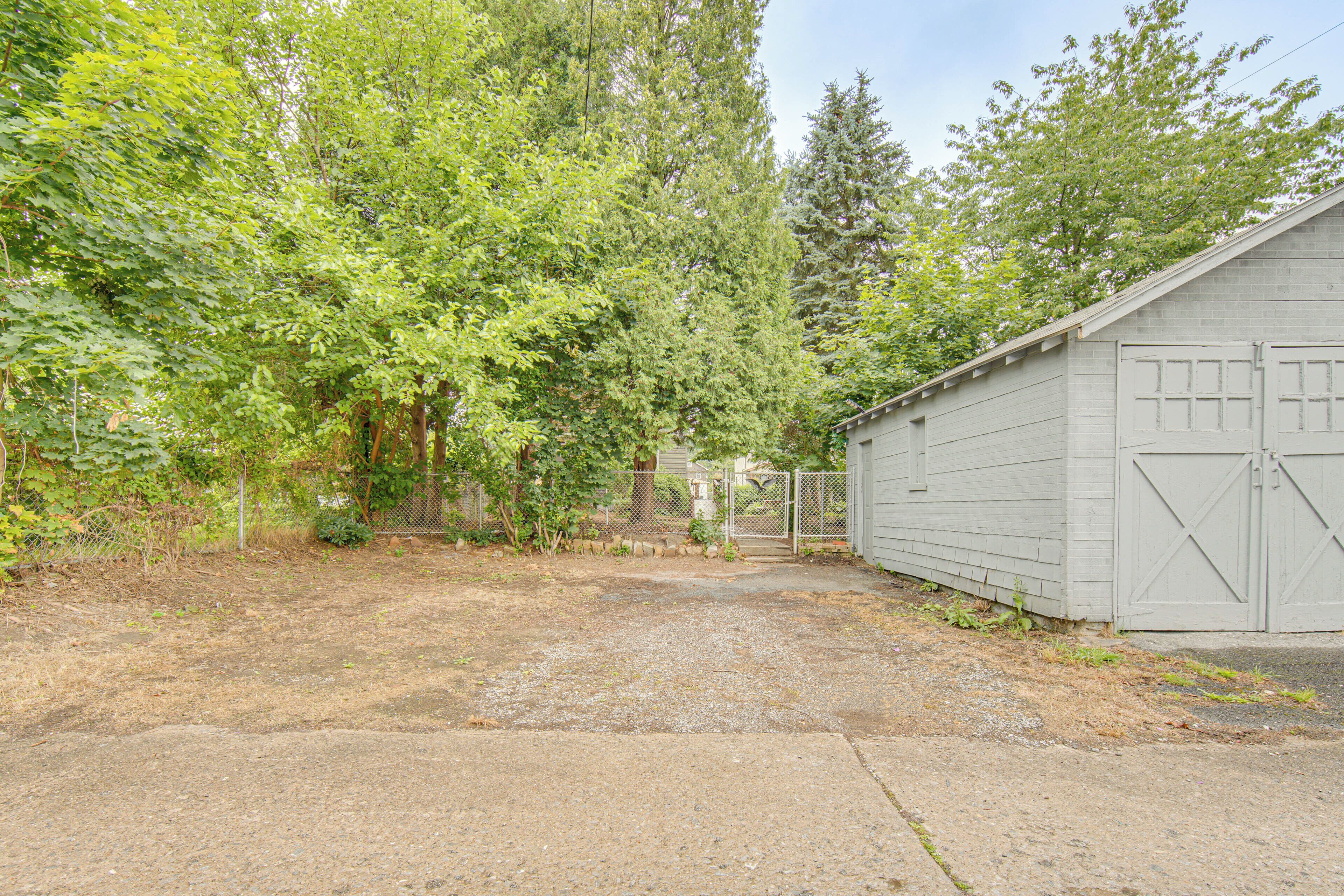 Une allée de gravier mène à un garage doté d’un portail en métal, le tout étant entouré d’arbres et d’une clôture.