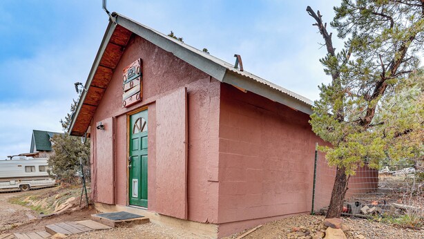 A small, single-story building with a red exterior and a green door, surrounded by a gravel driveway and a fenced area with trees. - Couples Haven 1 Mi to Dtwn Dolores! Bring Your Pet (Dolores)