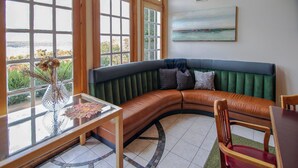 A room with a curved green sofa, a wooden table with a vase, and a view through large windows. - Hot Tub, Lake Monroe Views! 22-acre Outdoor Oasis (Bloomington)