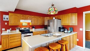 A kitchen with wooden cabinets, a white countertop, a stainless steel refrigerator, and a bar stool area. - Hot Tub, Lake Monroe Views! 22-acre Outdoor Oasis (Bloomington)