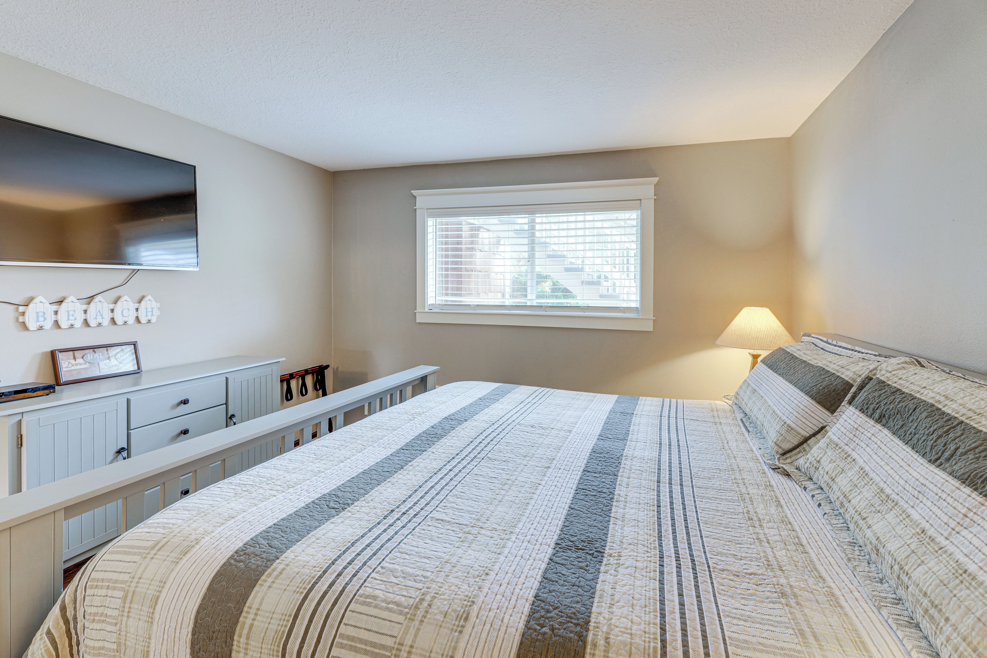 Une chambre à coucher comprenant un lit, une commode, un téléviseur fixé au mur, une fenêtre avec des stores et une lampe sur la table de chevet.