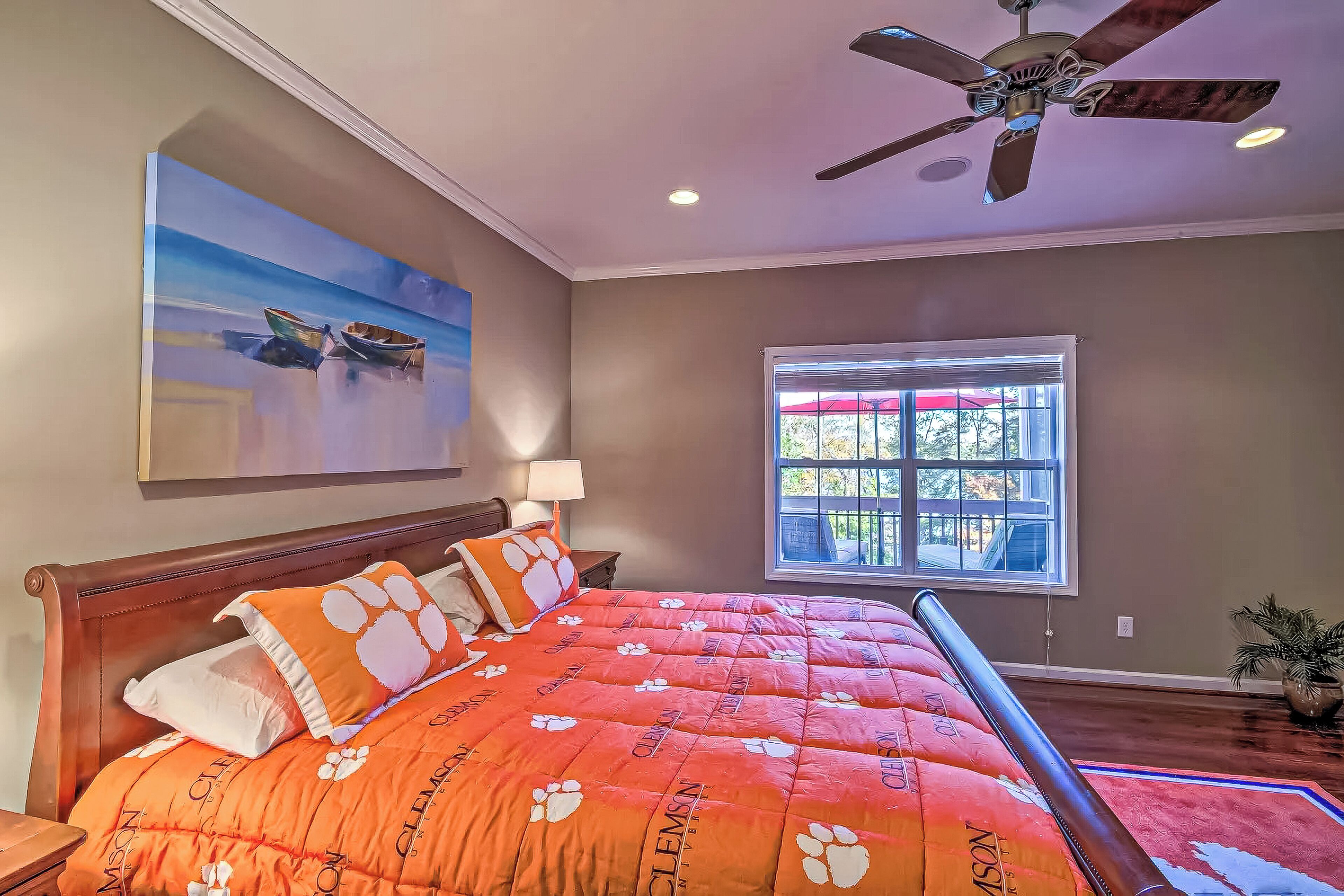 A bedroom with a large bed, a ceiling fan, a window with blinds, and a painting on the wall.