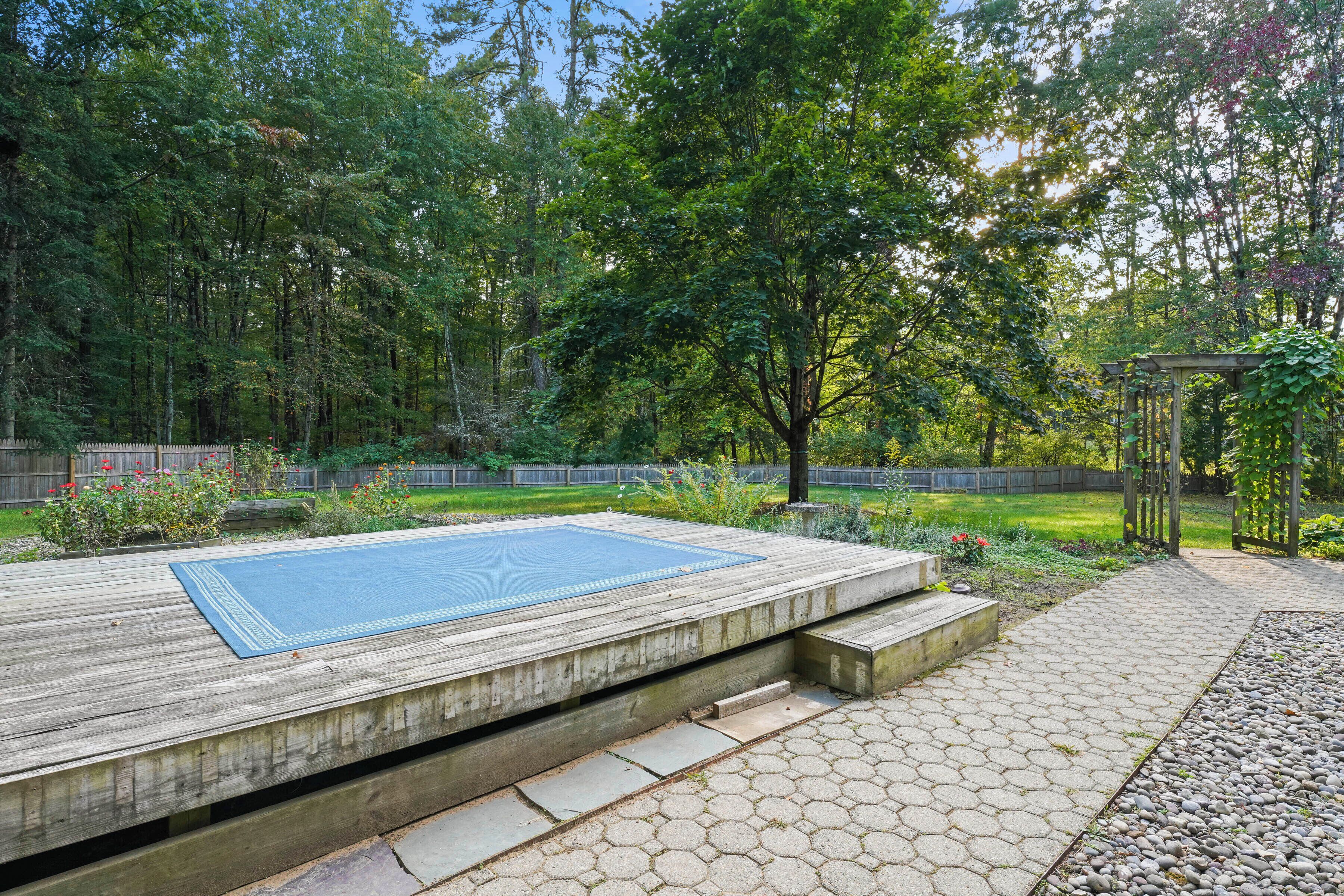 A wooden deck with a blue mat, hexagonal stone tiles, and a pergola with climbing plants.