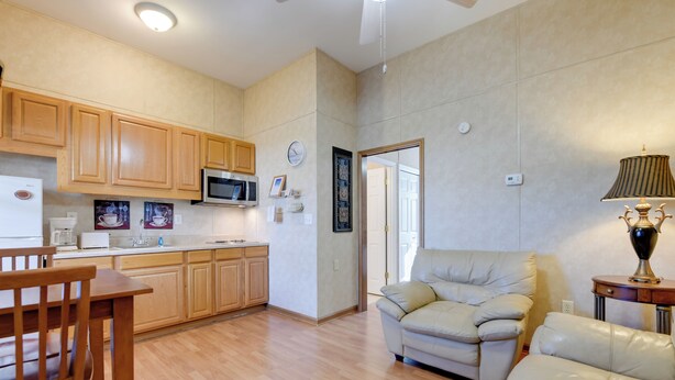 Una cocina moderna con armarios de madera, un microondas y un comedor con mesa y sillas de madera. Hay un sofá beige en la sala de estar y una lámpara sobre una mesita.