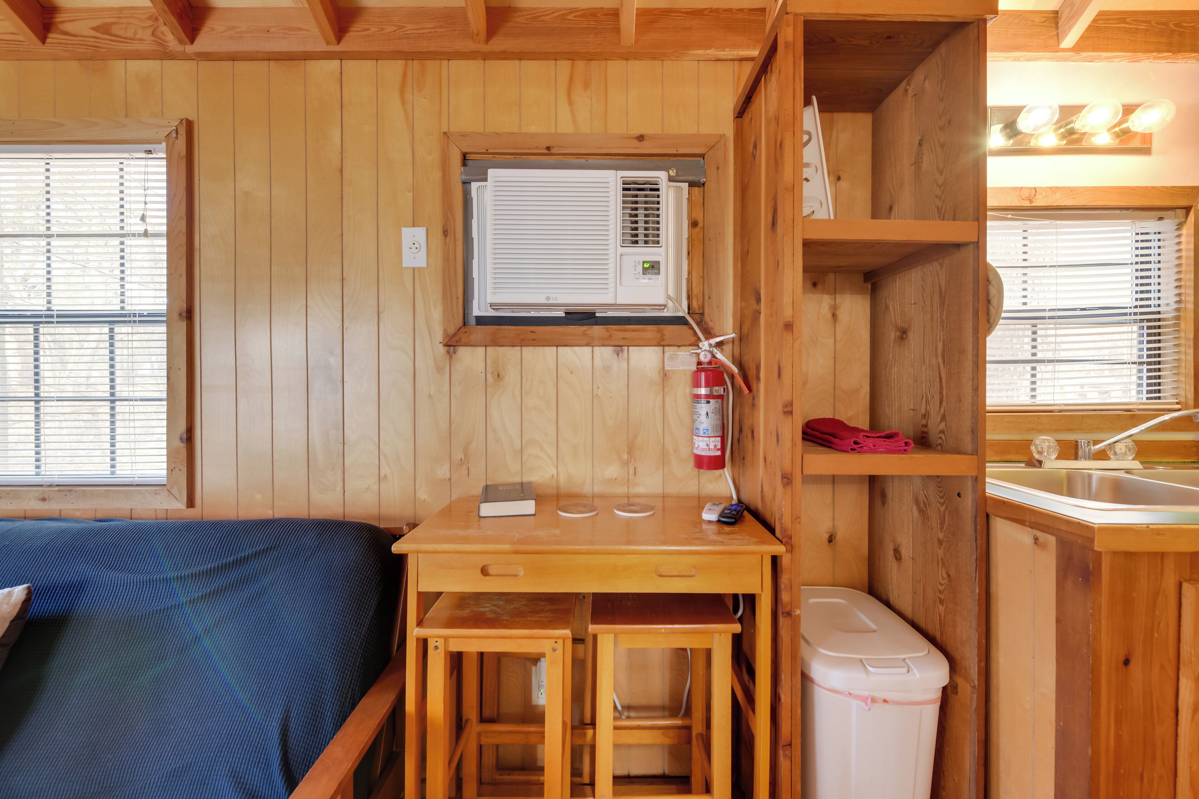 Una baita di legno con un letto, un piccolo tavolo con un libro, un estintore e una finestra.