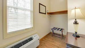 Une pièce avec une fenêtre, un climatiseur, un petit banc, une lampe et une étagère en bois.