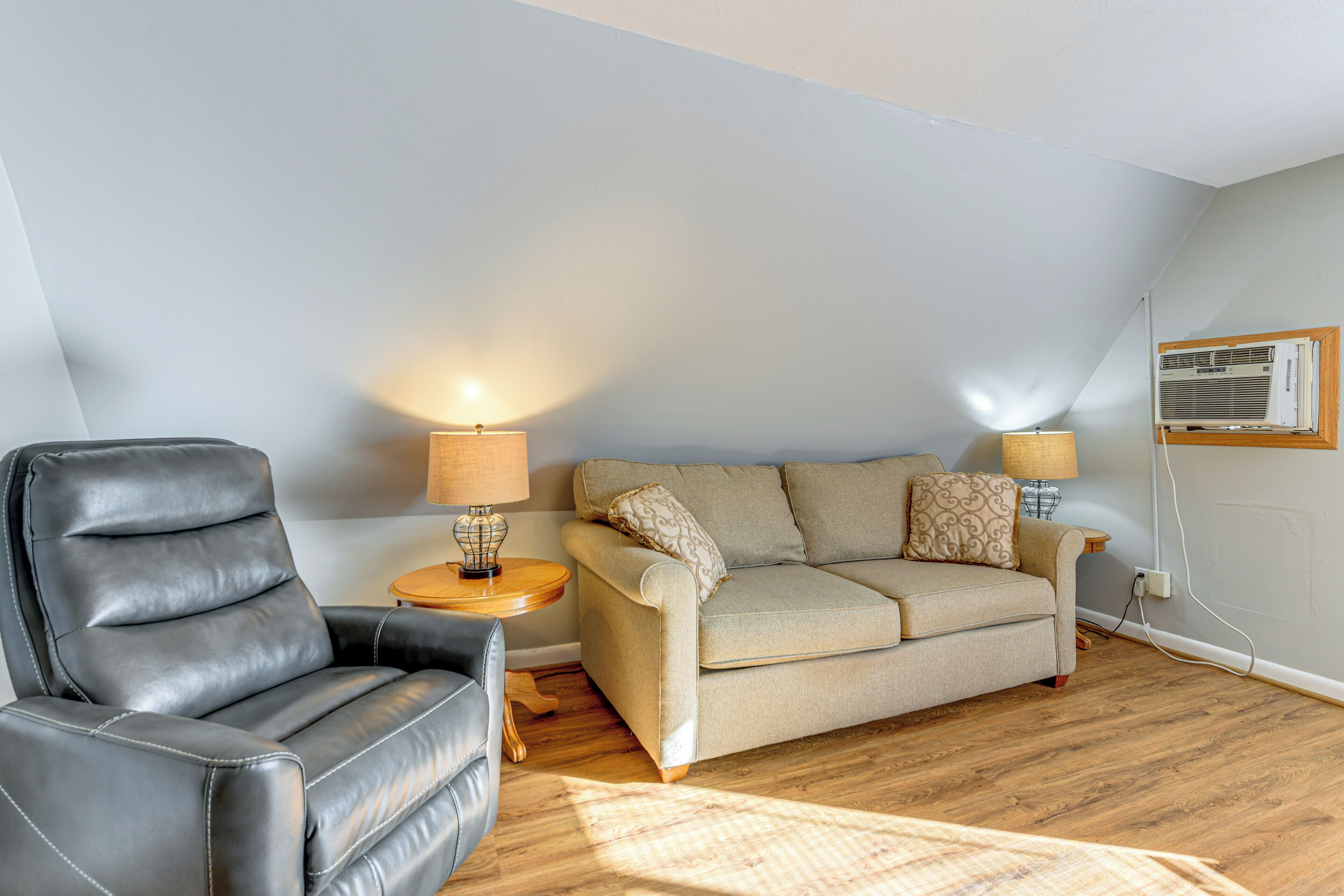 A living room with a leather armchair, a sofa, two lamps, and a wall-mounted air conditioner.
