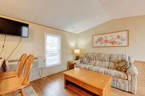 Ein Wohnzimmer mit einer Couch, einem Couchtisch, einem an der Wand montierten Fernseher, einem Fenster mit Jalousien und einem Gemälde an der Wand.