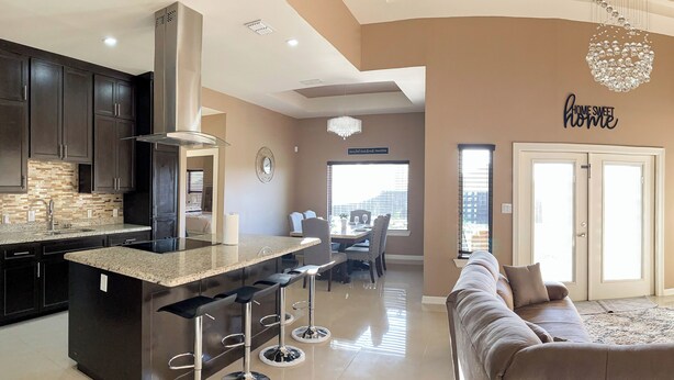 A modern kitchen with dark cabinets, a central island with bar stools, a chandelier, and a dining area with a sofa and chairs. - Pool, Patio & Yard: Chic South Texas Home (Olmito)