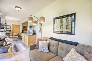 A living room with a sofa, a coffee table, and a dining area with a table and chairs. There is a kitchen area with cabinets and a refrigerator. - Sunny Downtown Studio w/ Kitchenette & AC (Fairbanks)