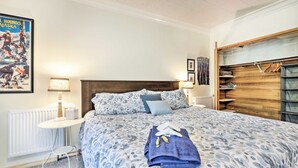 A bedroom with a patterned bedspread, a neatly made bed with towels, a bedside table with a lamp, a radiator, and a wooden wardrobe with hanging space. - Sunny Downtown Studio w/ Kitchenette & AC (Fairbanks)