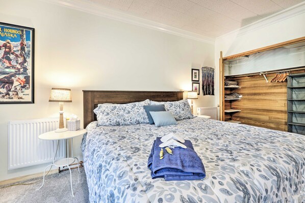 Un dormitorio con colcha estampada, cama bien hecha con toallas, mesita de noche con lámpara, radiador y armario de madera con espacio para colgar.