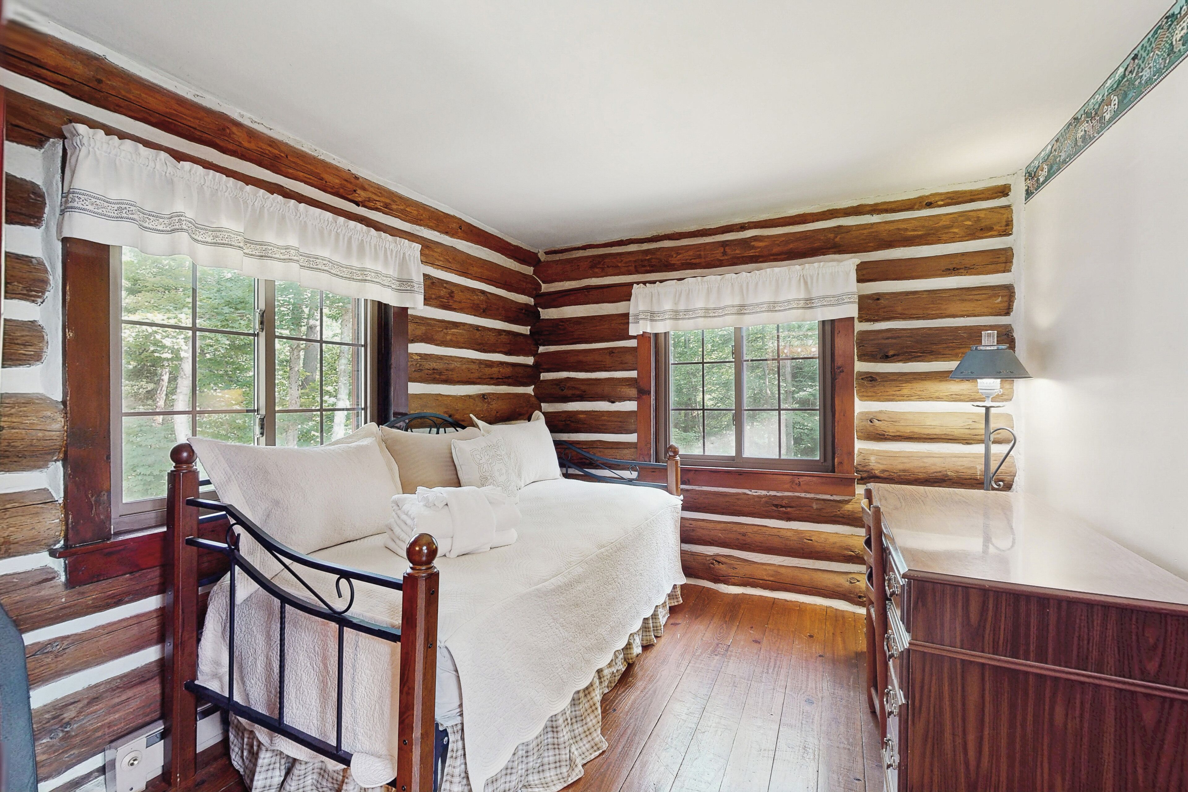 A log cabin bedroom with a wooden bed, two windows, a dresser, and a lamp.