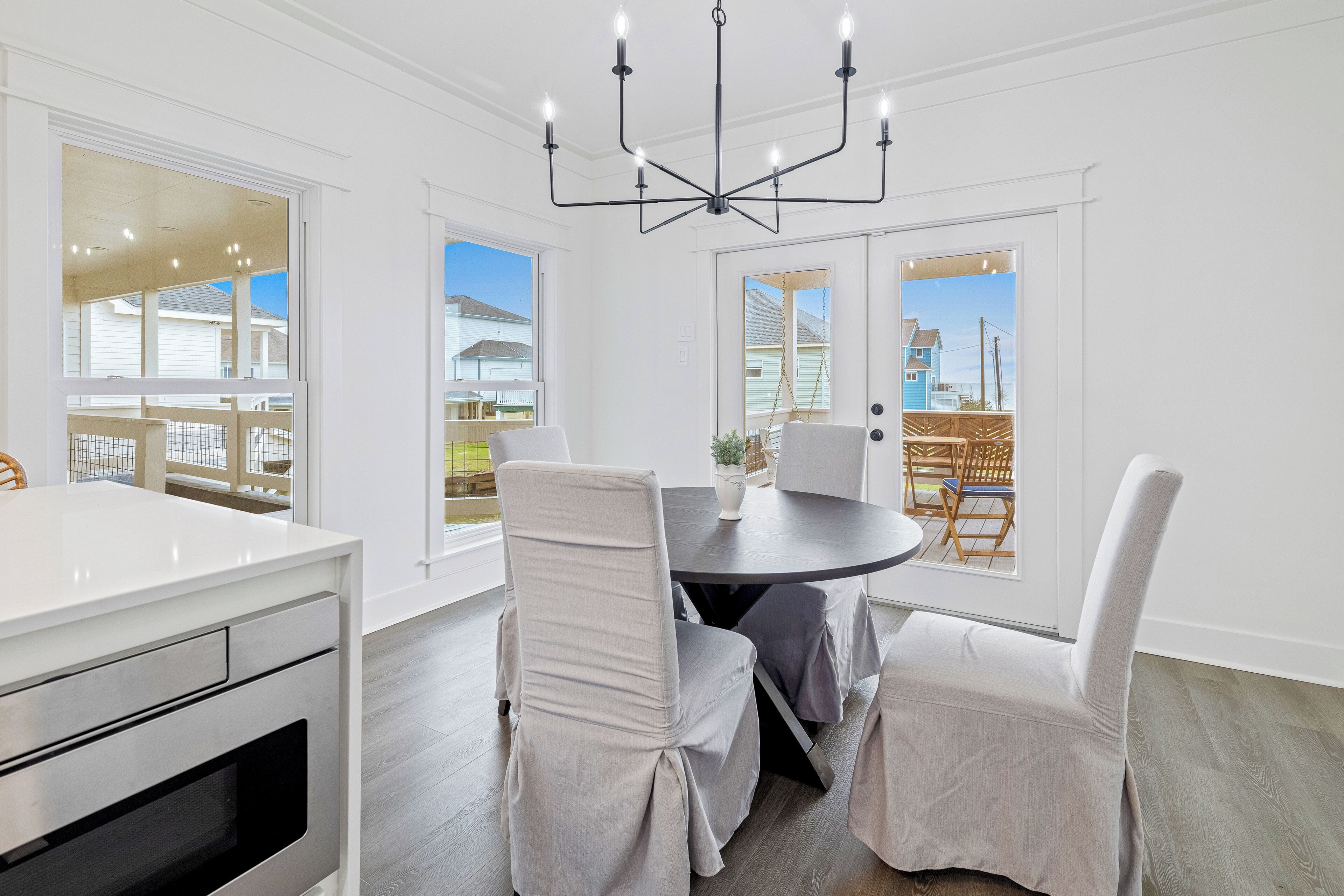 A dining area with a round table, four chairs, a chandelier, and large windows offering views of the outside.