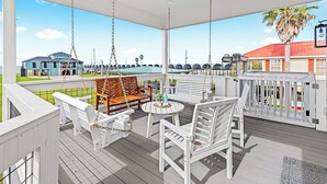 A covered outdoor deck with white furniture, including chairs and a table, overlooking a marina with boats and a building with a red roof. - Bring Your Boat! Lake Charles Home w/ Dock & Lift (Lake Charles)