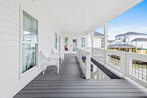 A covered wooden deck with white railings and large windows, leading to a covered porch area with white wicker chairs.