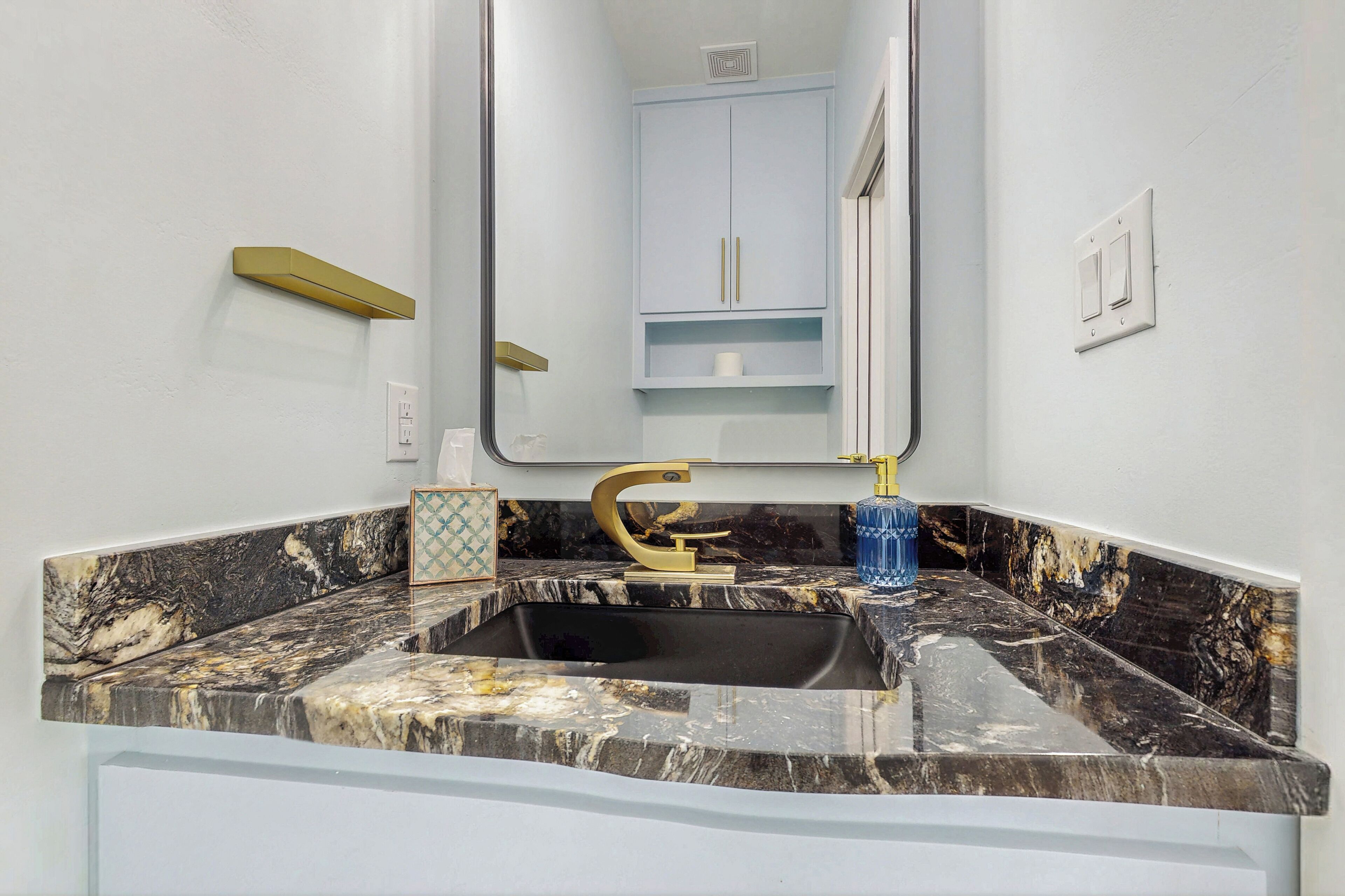 A bathroom with a marble sink, gold faucet, and a mirror reflecting a toilet and cabinet.