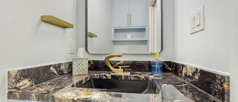 A bathroom with a marble sink, gold faucet, and a mirror reflecting a toilet and cabinet.