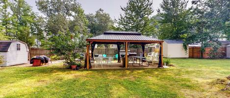 A covered patio area with a table and chairs, surrounded by a grassy yard and trees.