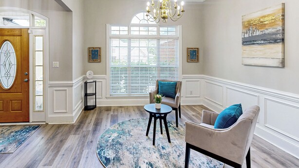 Une pièce avec une porte en bois, une grande fenêtre avec des stores, un lustre, un tapis rond, une petite table avec une plante et deux fauteuils avec des coussins bleus.